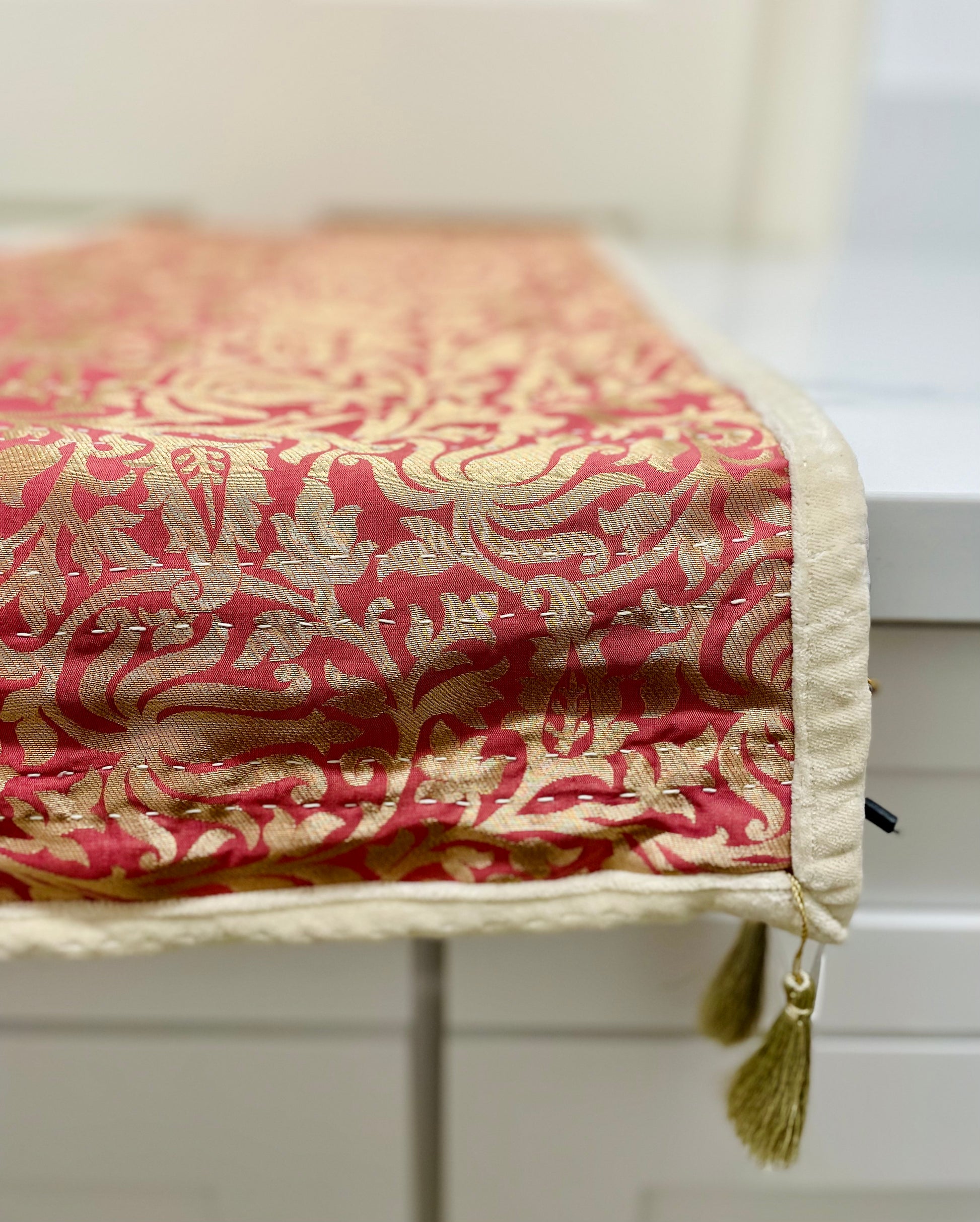 A close-up view of a elegant kantha table runner with a deep rose pink background and ornate gold detailing, finished with a beige velvet backing and adorned with golden tassels.